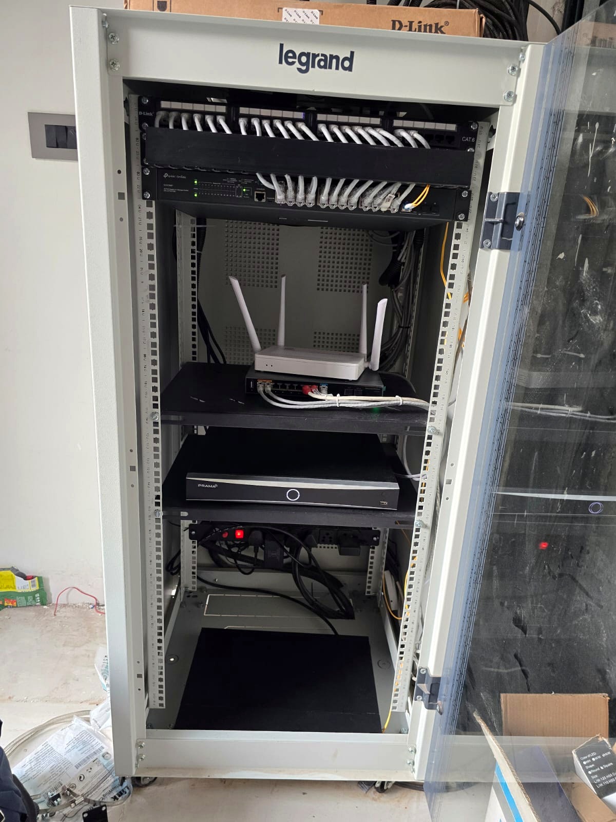 The Alibaug distribution rack: a Legrand floor-standing enclosure holding a D-Link Cat6 patch panel above a TP-Link Omada PoE switch, with the router, a Prama recorder and power distribution on the shelves below.