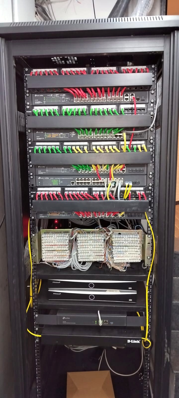 The Basilica Hotel server-room rack: colour-coded Cat6 patch leads landing on D-Link patch panels above TP-Link Omada switches, with punch-down telephony blocks, two Prama recorders and an Omada hardware controller mounted below.